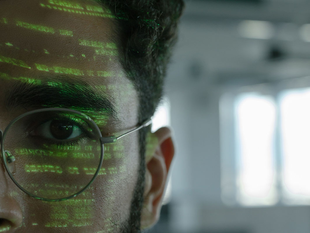 Mann mit Brille schaut in die Kamera, in seinem Gesicht spiegelt sich Code.