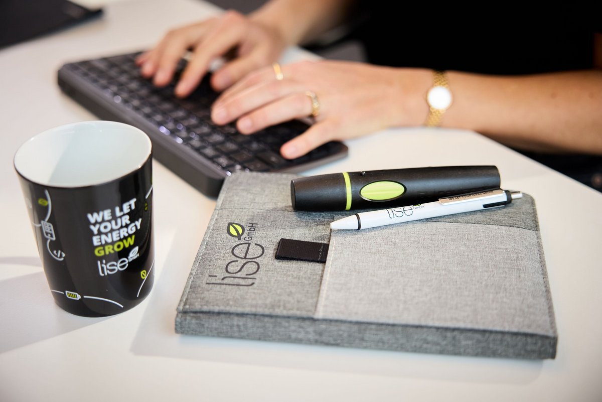 Ein lise-Entwickler tippt auf einer schwarzen Tastatur und vor ihm auf dem Tisch liegen eine Tasse, ein Notizbuch und Stifte