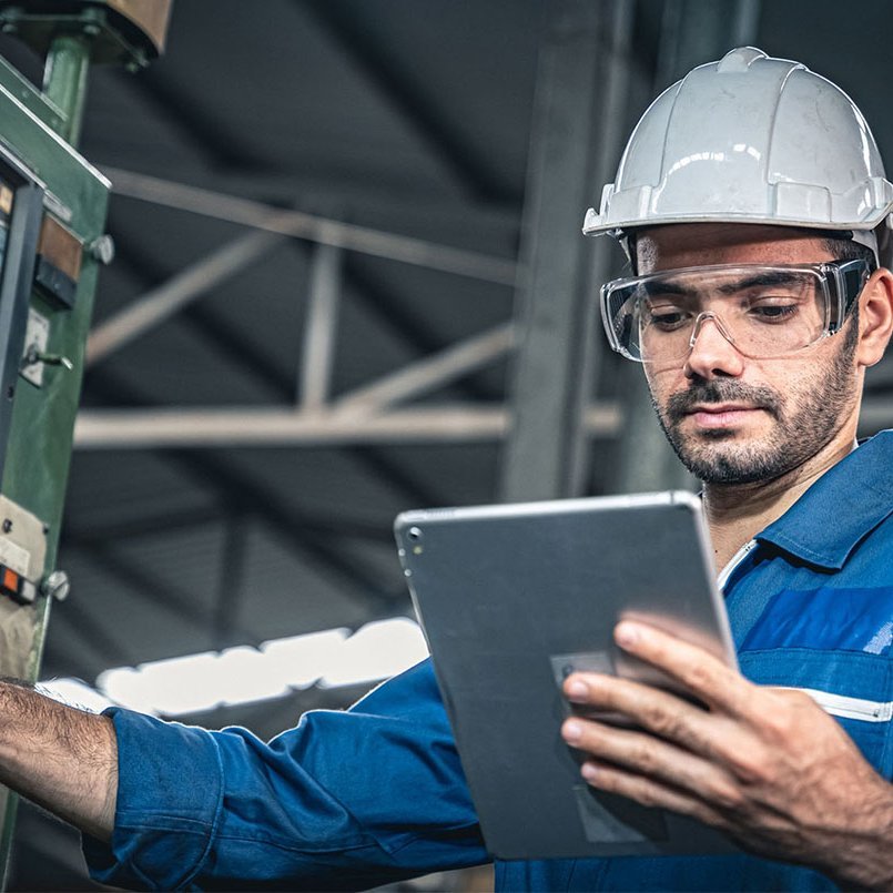 Industriearbeiter mit Schutzhelm bedient eine Maschine und nutzt dabei ein Tablet