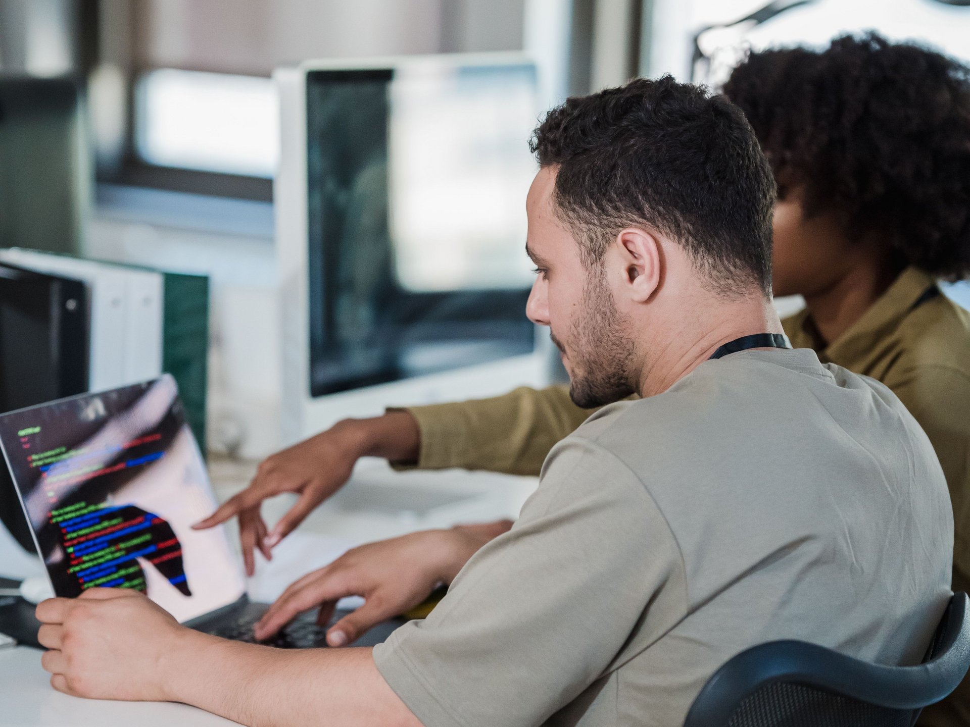 Zwei Personen sitzen am Schreibtischen vor einem Laptop und einem Monitor, eine Person zeigt auf eine Stelle im Code auf dem Laptopbildschirm.