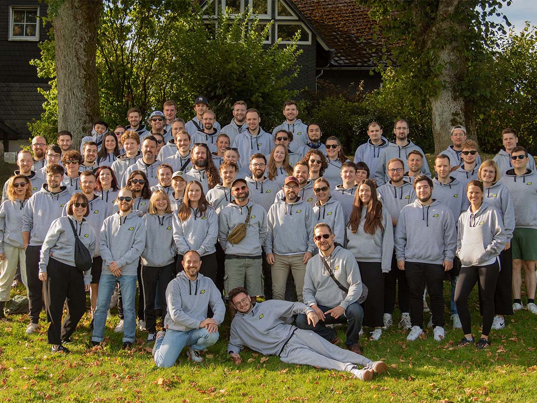 Ein Gruppenfoto der lise Mitarbeiter auf einer grünen Wiese. Sie tragen alle ihren grauen lise Hoodie. Im Hintergrund sind zwei Bäume vor einem Haus.