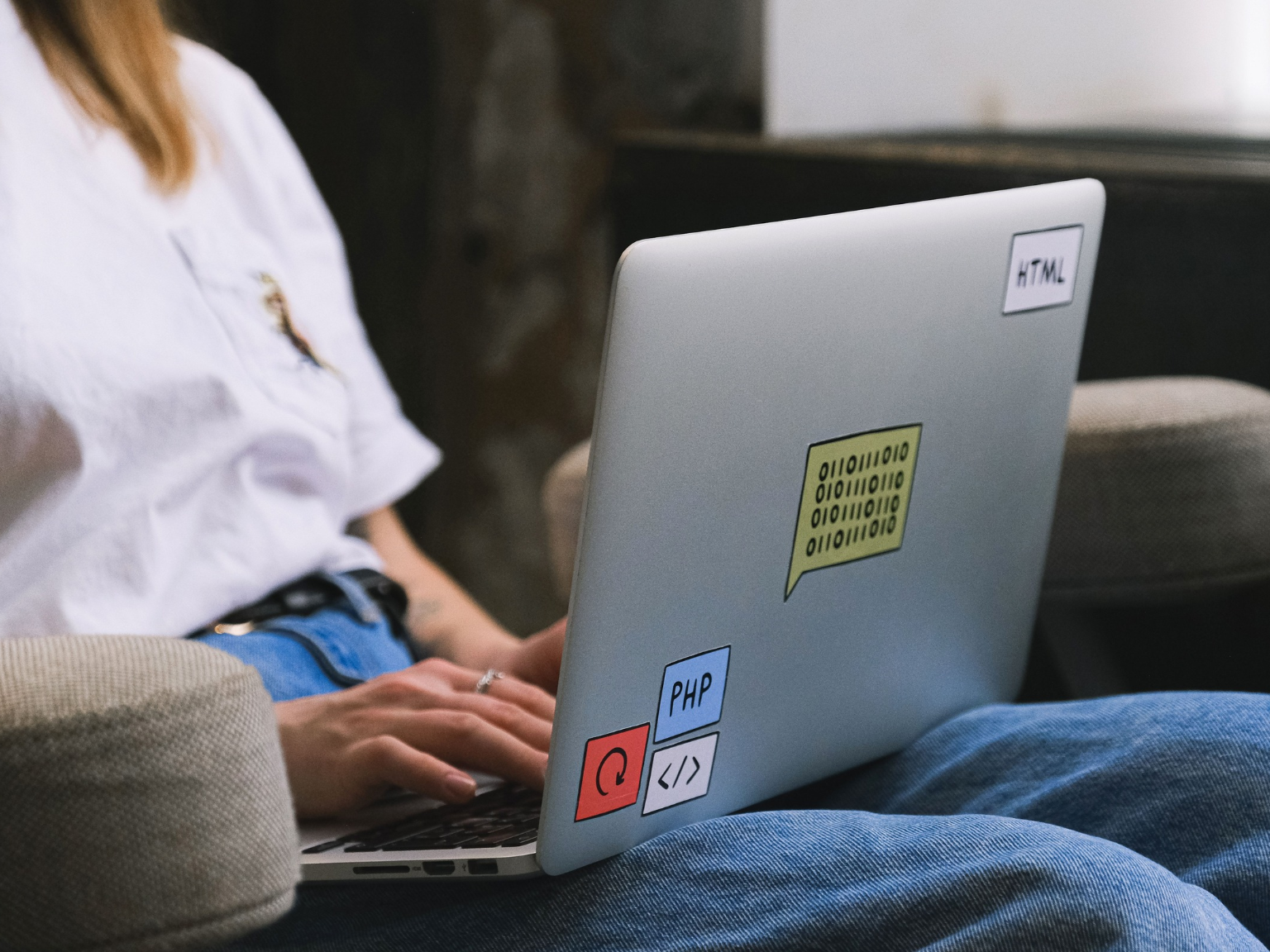  Person mit weißem T-Shirt und blauer Jeans sitzt auf einem Sessel und tippt auf einem Laptop mit mehreren Programmieraufklebern, darunter 'HTML’ und 'PHP'